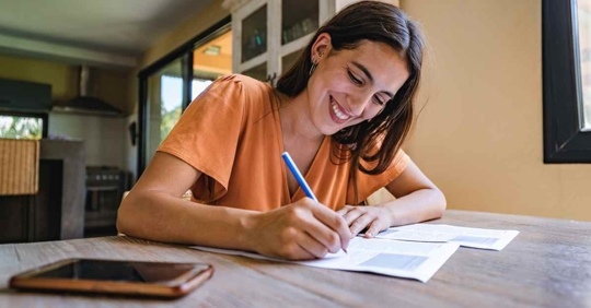 A woman writing something down while sitting at her kitchen table. She is smiling