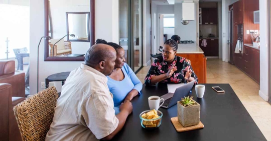 A man and woman sitting next to each other while another woman reviews something. They are all sitting at a kitchen table