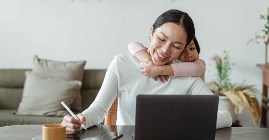 A woman working on something at her desk while her daughter hugs her from behind